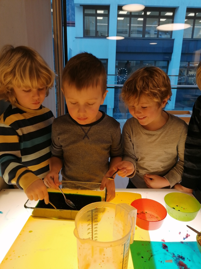 drei junge Buben experimentieren mit einem Löffel in einer Schüssel. Zwei sind sehr konzentriert auf die Arbeit, der andere lacht.