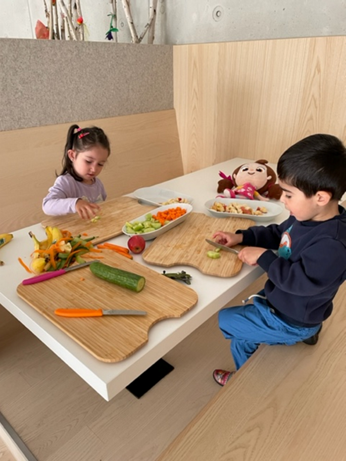 Ein kleiner Junge und Mädchen schneiden verschiedestes Obst auf, unter anderem Gurken, Äpfel, Karotten und einiges mehr.