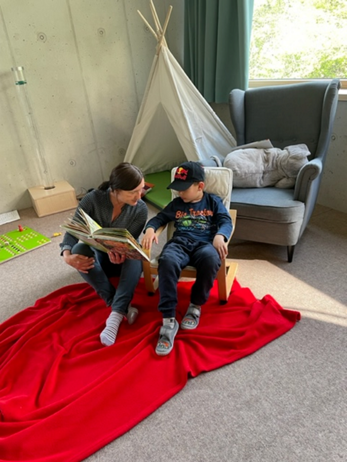 Eine Kindergartenpädagogin liest einem Junge ein Buch vor und zeigt im gerade etwas in diesem, vielleicht ein Bild zur Veranschaulichung.