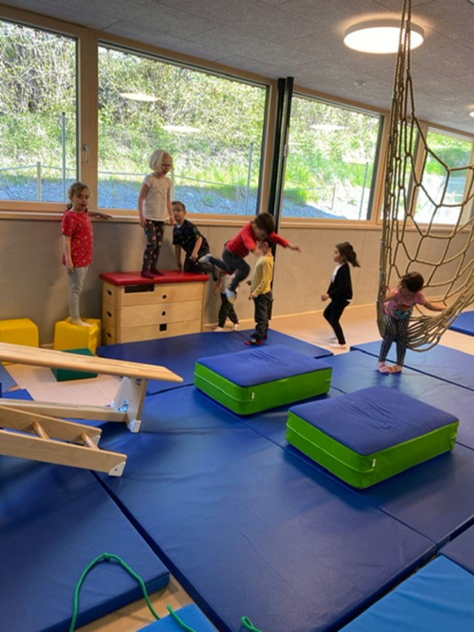 Viele Kinder haben sich in der Turnhalle einen Parkour aufgebaut und einer springt gerade von einem Kasten auf ein Kissen. Die nächste wartet schon auf dem Kasten um zu springen. Die anderen Kinder stehen auch schon hinter diesem Mädchen an, den Parkour zu bewältigen.